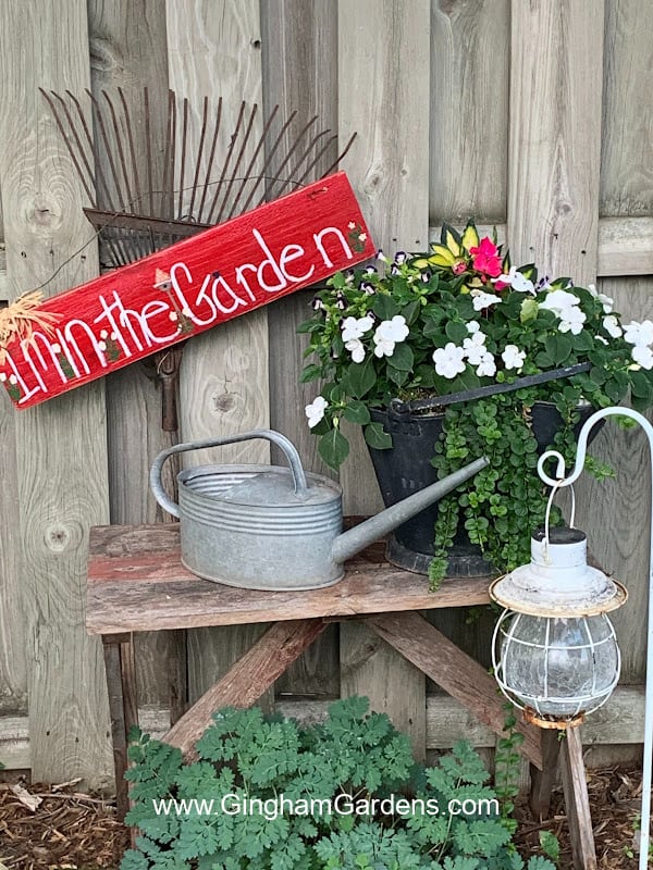 Cute grouping in a flower garden that includes a bench, a coal bucket with flowers, a watering can, a solar light, and an old rake with a garden sign hanging on it that says in the garden.
