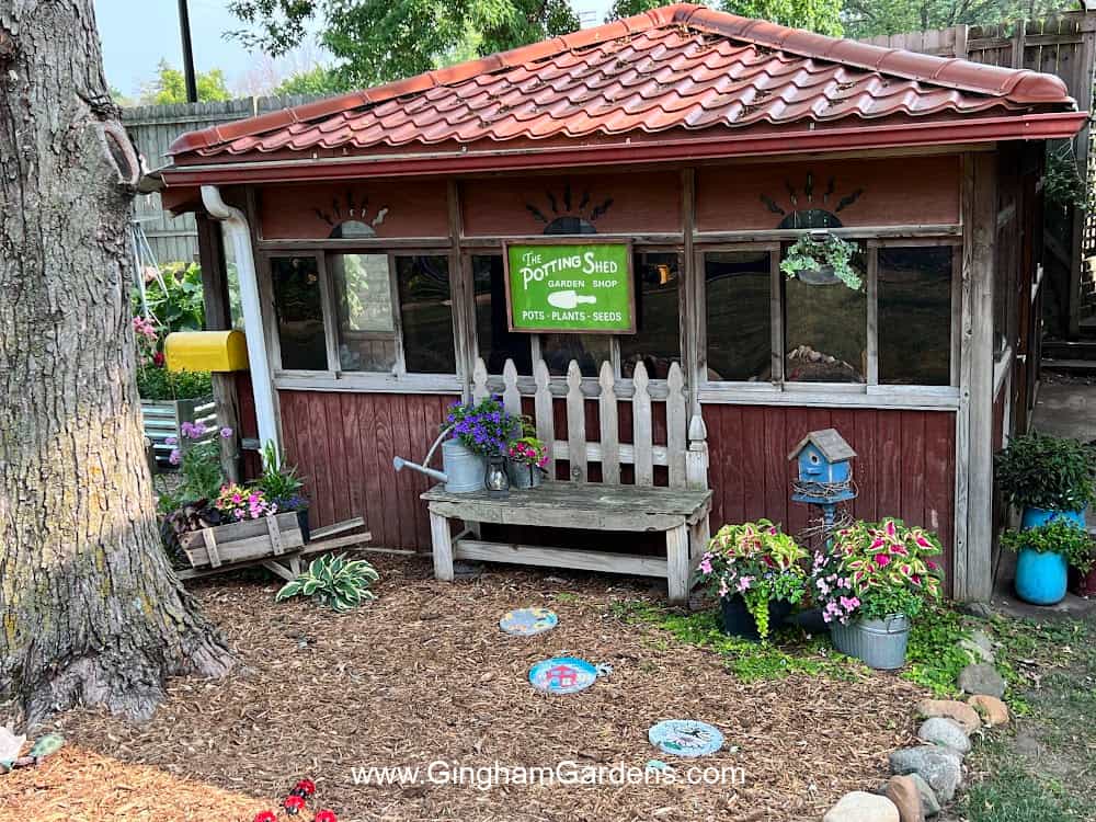 The outside of a potting shed with a sign and various pots of flowers and a bench.