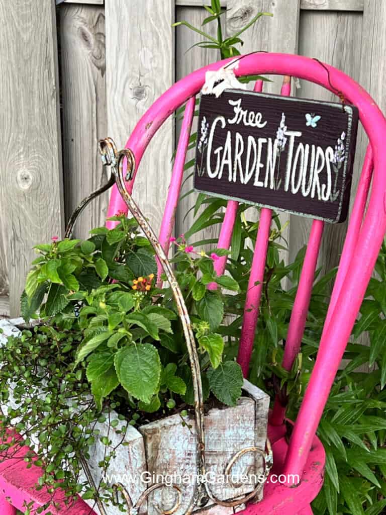 Bright pink chair in a garden with a planter of flowers on the seat and a 'garden tours' sign on the back.
