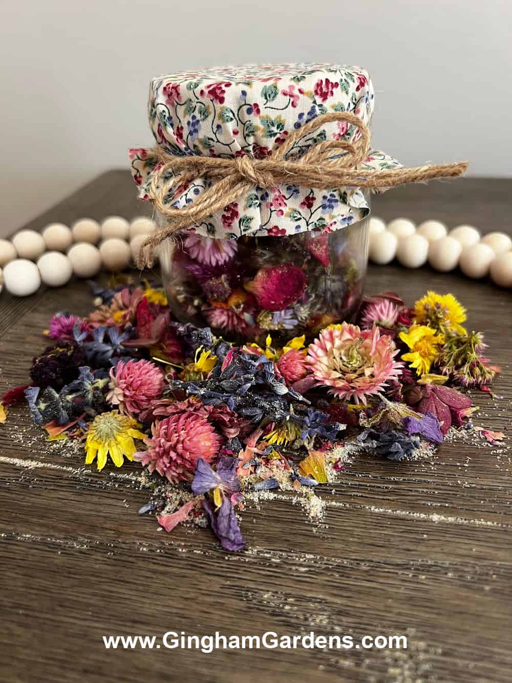 A jar of potpourri with a fabric cover tied with jute surrounded by colorful potpourri.
