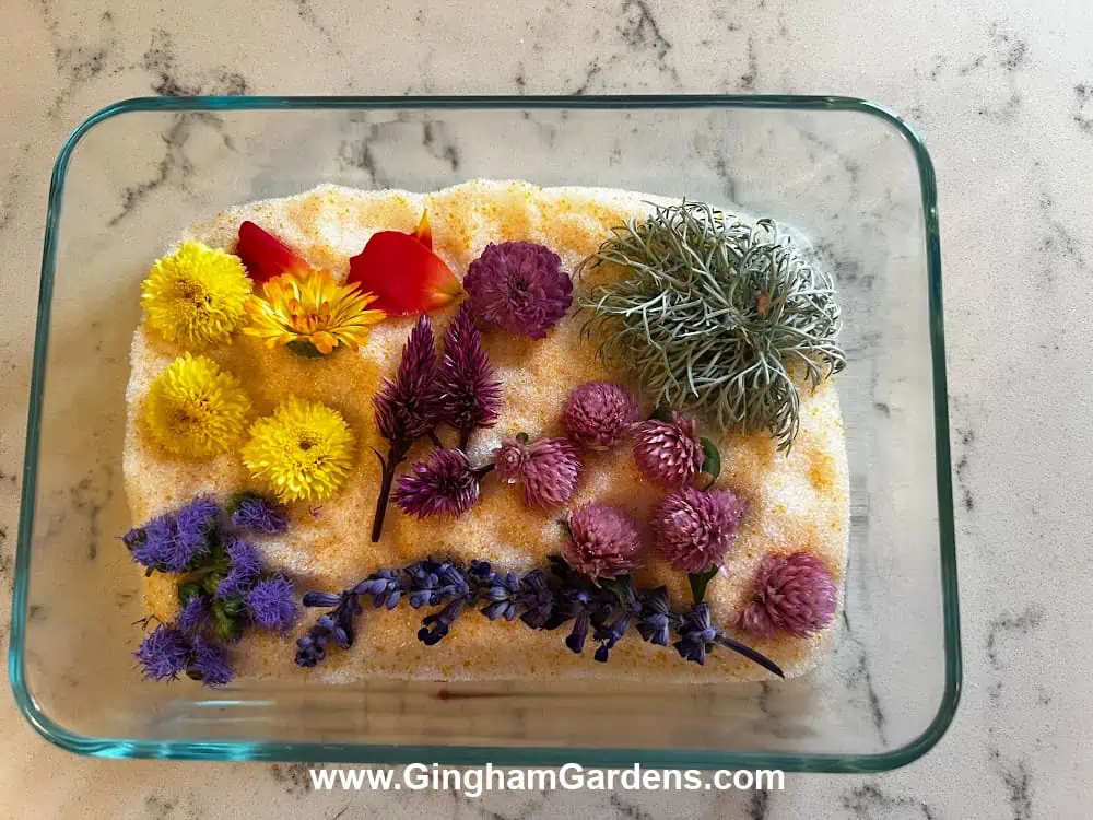 Glass baking dish with silica gel granules and flower heads layered on top for making homemade potpourri.